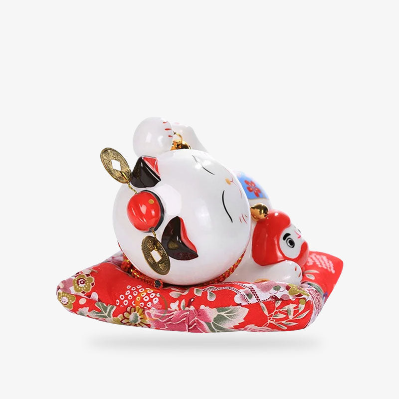 Un chat maneki neko ceramique allongé sur un coussin rouge et qui tient dans sa patte droite une statuette rouge