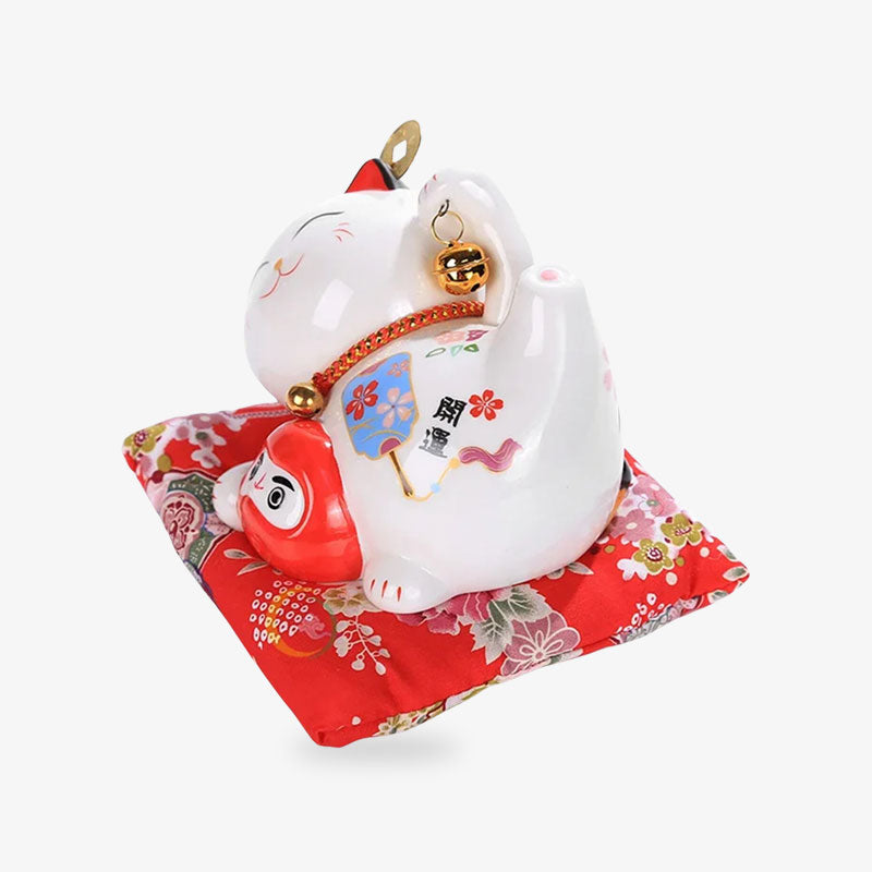 Ce chat maneki neko japonais ceramique est une statuette porte-bonheur japonais. Le porte-bonheur est allongé sur un coussin rouge et tien dans sa patte un Daruma Rouge