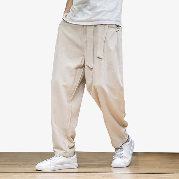 Un homme porte un pantalon style japonais de couleur beige. Il aux pieds une paire de sneakers blanche. Le pantalon est large avec une ceinture