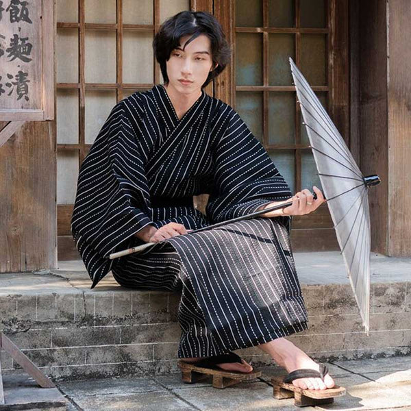 Un japonais est habillé avec un kimono homme noir. Il tient dans la main une ombrelle japonaise, et a enfilé des sandales en bois geta