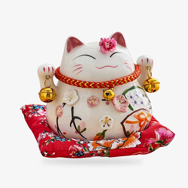 Ce Maneki neko porte bonheur chat japonais est posé sur un coussin rouge. La statuette porte-bonheur lève les deux pattes et tient deux clochettes. C'est un objet de décoration japonaise posé sur un coussin rouge
