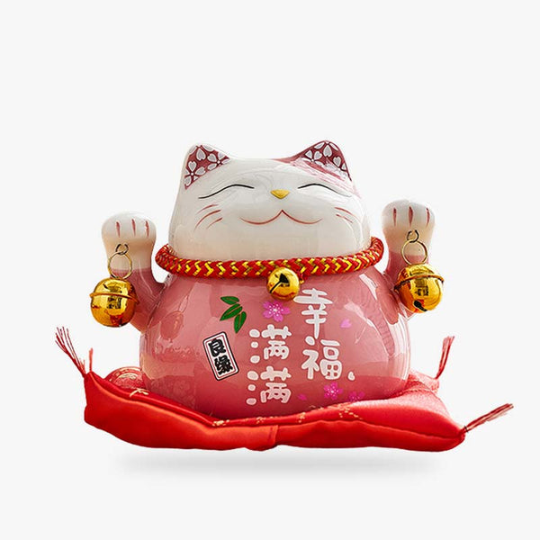 Une tirelire Maneki neko de couleur rose. Le chat japonais a les deux pates levées et tient des clochettes dorés. Des kanji sont peints sur le corps de la statuette japoanise. Cet objet décoration japonais est posé sur un coussin rouge