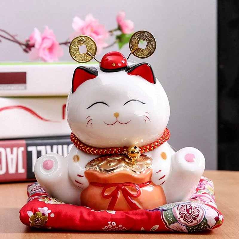 LE chat japonais porte-bonheur-céramique est sur un coussin rouge. Il a un sac rempli d'argent