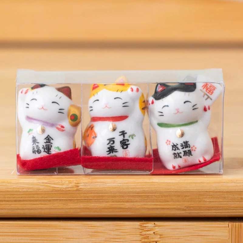 trois petits chat porte-bonheur blanc sont posés sur une tables. Les chats maneki neko sont emballés dans une boite transparente