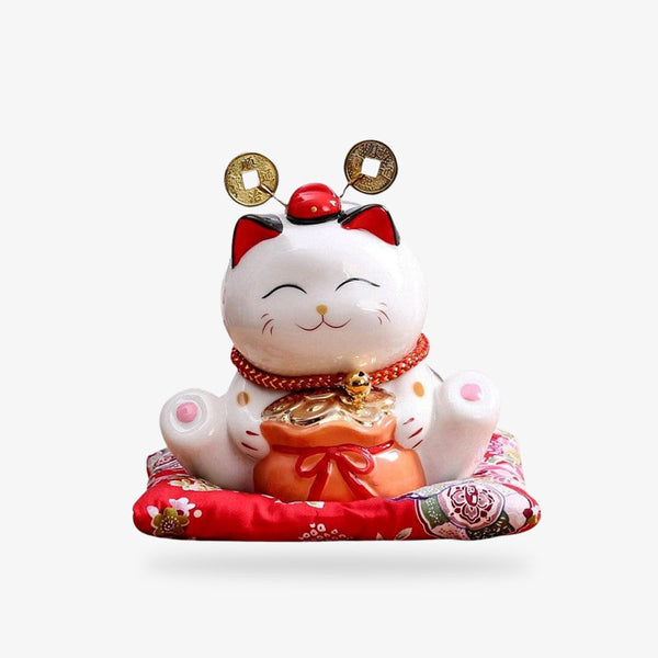 Ce chat porte bonheur Japon est de couleur blanche. Il est en ceréramique sur un coussin rouge. Il a un sac rempli de pièces d'or