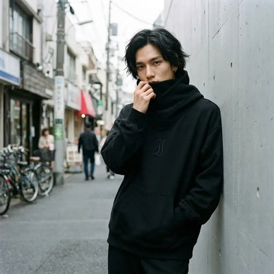 Hoodie streetwear noir de Tokyo porté avec le col cheminée remonté sur le bas du visage, contre un mur