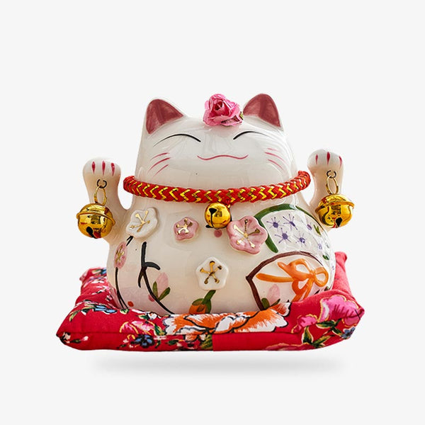 ce maneki neko porte-bonheur est une statuette de chat japonais de couleur blanche et en céramique. Le chat porte-bonheur tient dans chacune de ses pattes des clochettes. Cet objet deco est positionné sur un coussin rouge