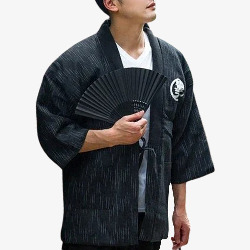 Uh homme est habillé avec un manteau japonais hanten de couleur noir. La matière en coton est molletonné. Le tissu est imprimé avec un kanji blanc. L'homme tient dans la main un éventail pliant japonais noir pour compléter sa tenue