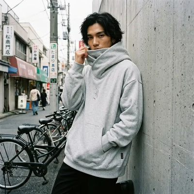 Sweat oversize Tokyo porté avec le col cheminée remonté sur le bas du visage, devant un mur en béton.