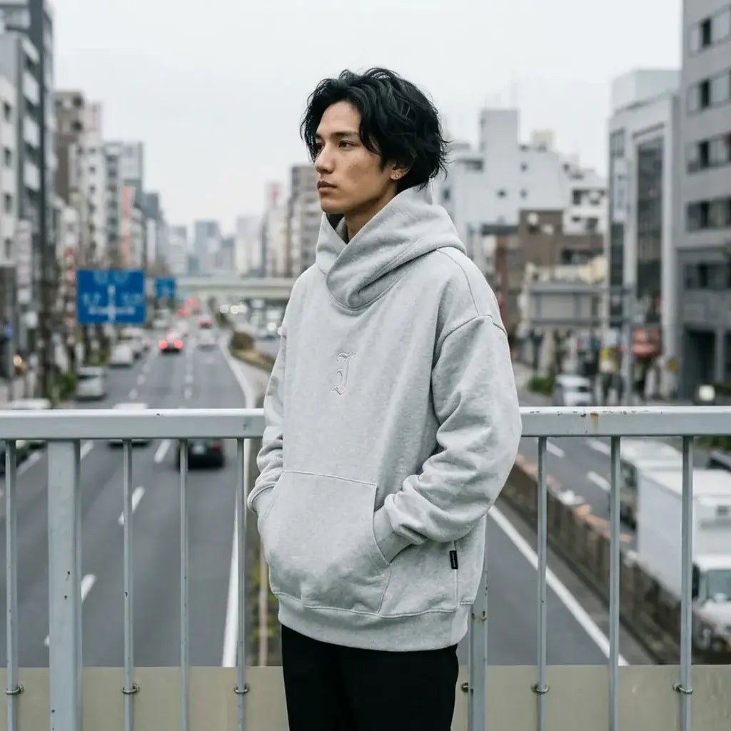 Sweat à capuche gris streetwear japonais porté sur une passerelle piétonne au-dessus d'une large avenue de Tokyo, sous un ciel couvert.