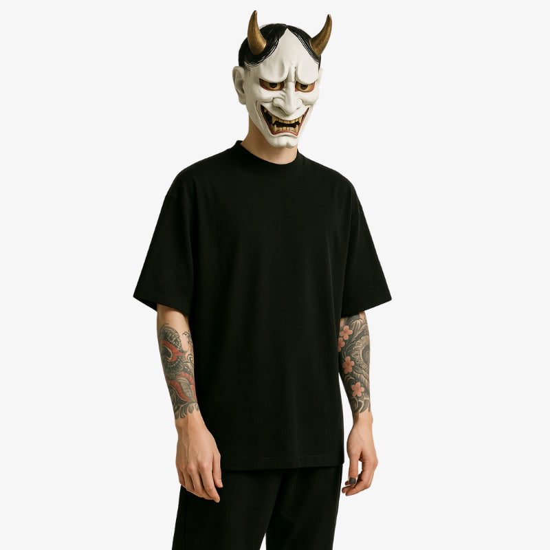 Un homme est vêtu d'un t shirt harajuku noir. Son visage est dissimulé par un masque blanc Hannya. Il porte des tatouages irezumi sur les bras. Le t-shirt japonais est oversize.