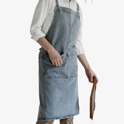 Tablier en coton denim épais bleu, avec bretelles croisées et poche avant pratique pour un usage polyvalent.