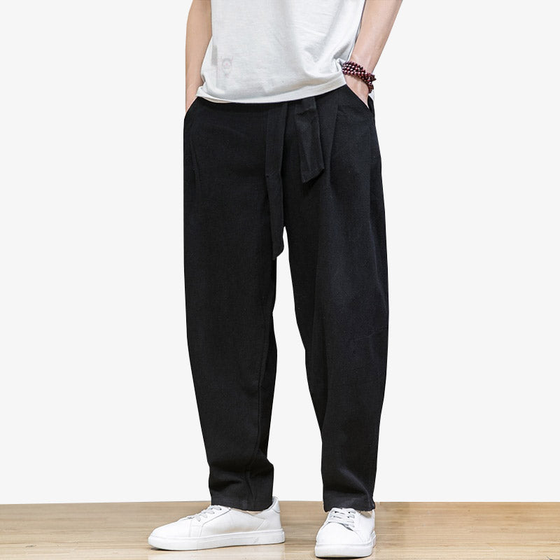 Cet habit large noir est un pantalon homme style japonais avec une ceinture pour l'attacher. L'homme est debout, il porte des chaussures basses blanche et un bracelet en perle